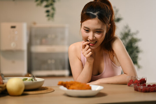 Young Asian Woman Tempted by Fried Chicken While on Diet, Sitting at Kitchen Table with Healthy Foods