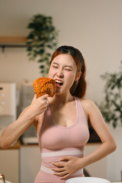 Young Asian Woman in Workout Clothes Eating Tempting Fried Chicken While Holding Belly Indoors