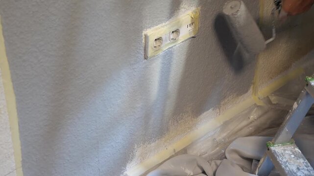 Professional Painter Rolling Grey Paint on Lower Wall Around Power Outlets Standing Near Stepladder Home Renovation Precision DIY Project Indoors