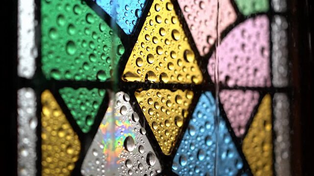 Water droplets cling to a multi colored stained glass window, light passes through yellow, blue, green and pink triangular glass panels, dark frames separate sections