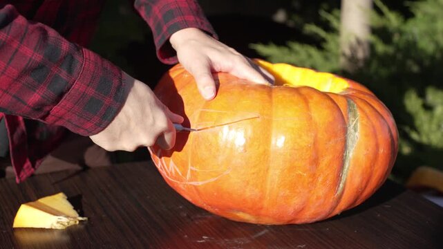 Close up of hands carving scary eye socket pushing out ripe squash flesh using kitchen knife in golden hour, authentic autumn holiday craft