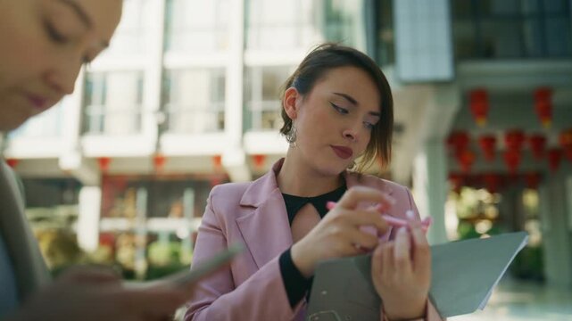 Two professional businesswomen collaborate outdoors, reviewing documents and discussing project details. They are actively engaged in brainstorming new ideas and taking important notes.
