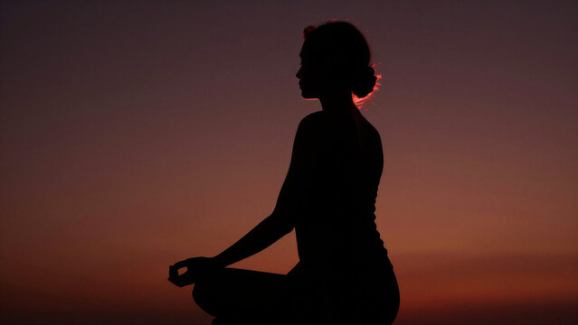 Profile silhouette of woman meditating in zazen pose with deep red sunset background, copy space on the left, diagonal back view