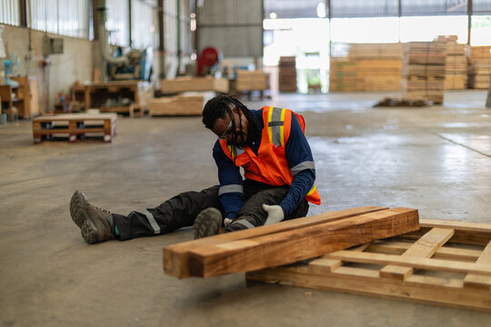 Nursing an injury after a workplace accident, a seated worker holds his leg beside stacked timber and a wooden pallet.