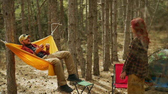 Couple toss an apple before sharing downtime in a hammock at a sunny forest campsite while camping.