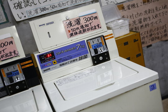 A commercial SANYO top-loading coin washer in a laundromat features a detergent dispenser and price signage.
