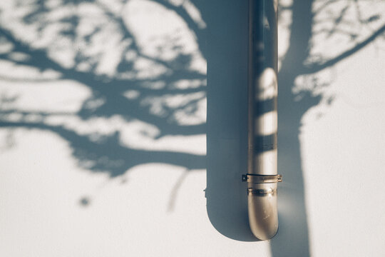Metallic downspout pipe mounted on a white wall with soft tree branch shadows cast across the surface.