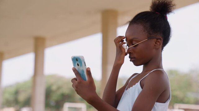 Child uses digital devices in everyday settings. Girl interacts with her phone while putting on eyeglasses outdoors under covered area smartphone glasses