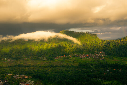 Aerial view of the lush green hills and rice terraces with low-hanging clouds clinging to the mountainside under golden sunset light in Sidemen, Bali, Indonesia.