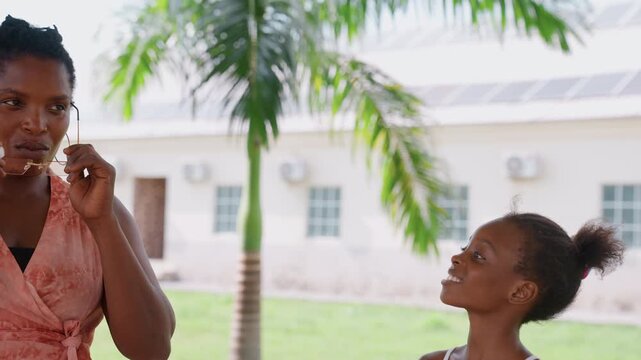 Woman in pink interacts casually outside near palm trees. Woman holds eyeglasses while conversing with child. building and greenery form background details