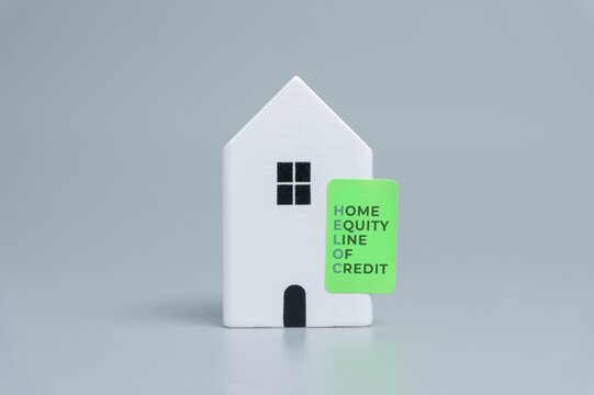 White miniature house with a green label reading HOME EQUITY LINE OF CREDIT on a gray background. Property financing, secured borrowing, mortgage lending, homeowner debt, residential equity concepts.