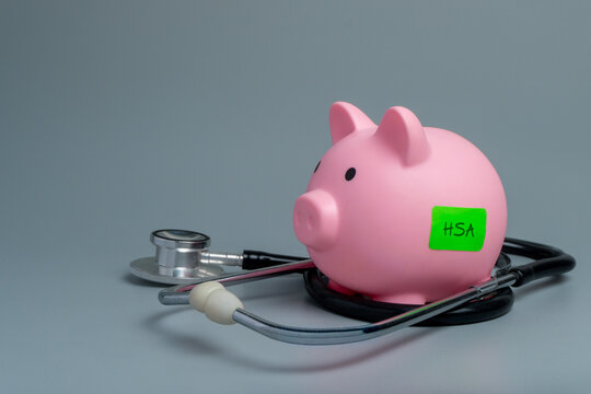 Pink piggy bank labeled HSA surrounded by a stethoscope on a gray background. Health savings, medical expenses, healthcare finance, insurance planning concept.
