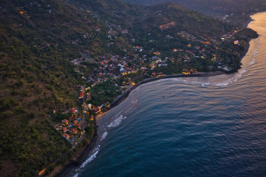 Aerial view of Lipah Beach at twilight, featuring the coastal village with warm glowing lights, lush green hills, and the dark sea along the shoreline in Kecamatan Abang, Bali, Indonesia.
