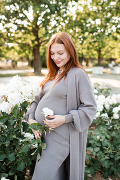 Smiling cute pregnant woman 24 - 29 year old wearing maternity clothes with rose flowers in garden outdoors. Prenatal lifestyle.