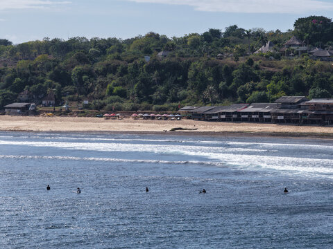Aerial view of Balangan Beach with surfers in the water, sandy coastline, beach huts, and lush green hills in the background in South Kuta, Bali, Indonesia.