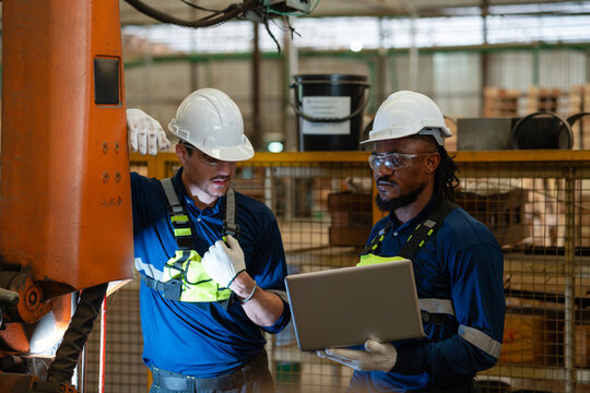 Orange industrial robot under inspection for automation stands beside two men sharing a laptop inside a fenced cell.