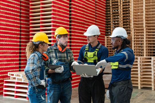 Pallets surround factory workers practicing teamwork during a team meeting as four colleagues discuss a laptop and a tablet.