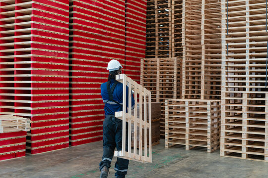A wooden pallet used for warehouse logistics is carried on a worker's shoulder beside tall stacks of wooden pallets.