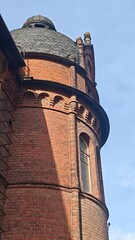 Red brick building with domed slate roof, ornate masonry, arched windows, decorative chimneys, and curved architectural facade against blue sky in historic architecture © Olena