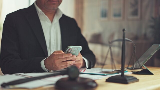Mature lawyer texting on mobile phone at office desk beside gavel scale. Legal advisory consultation, corporate compliance management, digital court case research, attorney communication.