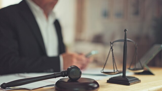 Wooden gavel balance scale rest on desk before male lawyer working in background. Legal advice consultation, judicial trial hearing, contract litigation courtroom justice concept.