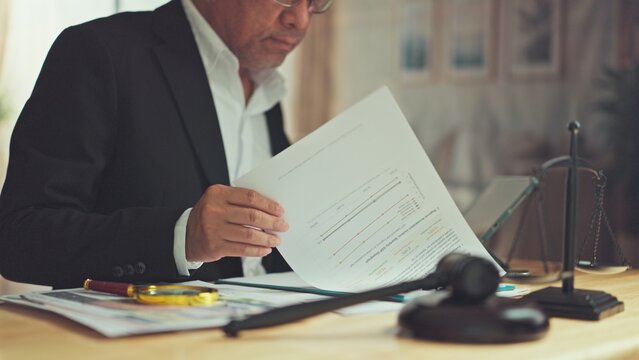 Mature lawyer examines financial report papers beside gavel scale desk. Corporate fraud investigation audit, courtroom evidence review, commercial litigation analysis.