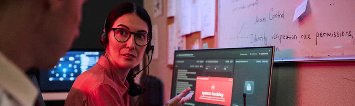 Caucasian middle aged woman wearing headset discussing cyber threat alert on computer monitor with male colleague. Image suitable for IT security, data defense, corporate software marketing