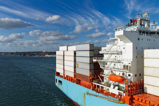 Aerial view of a large container ship with stacked cargo containers sailing on the sea under a blue sky with clouds near the coast of the United Kingdom.