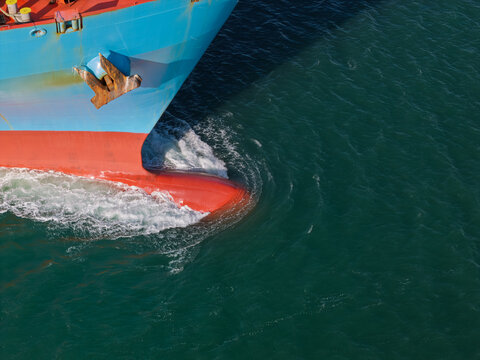 Aerial view of the bow of a large container ship with a blue and red hull cutting through deep green water and creating a bow wave in the United Kingdom.
