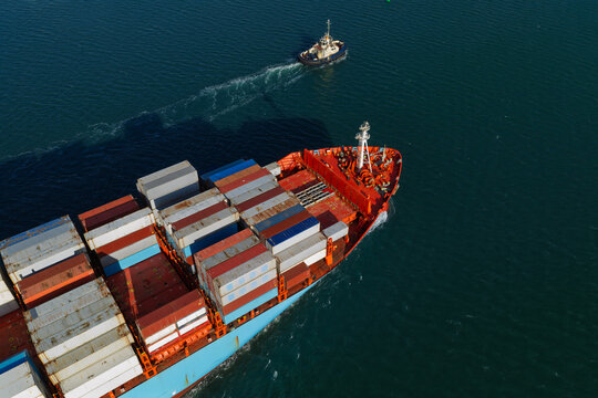 Aerial view of a large container ship loaded with colorful shipping containers being guided by a tugboat through deep blue sea waters in the United Kingdom.