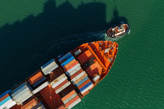 Aerial view of a large container ship being guided by a small tugboat through green sea waters in the United Kingdom.