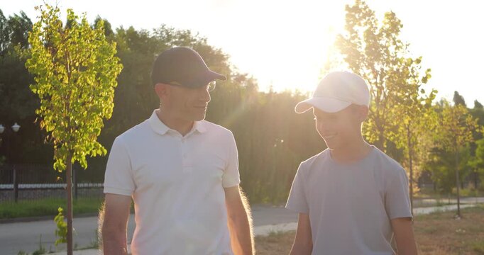 Father and son walk through park and talk together at sunset. They share warm family conversation in summer evening light. Outdoor bonding supports parenting, family, and companionship stories.