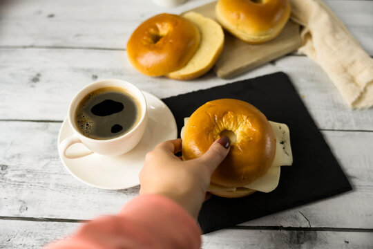  Morning coffee cup with fresh bagel and swiss cheese slice on white wooden table/&bull; A high-angle horizontal shot of a delicious breakfast setup on a rustic white wooden table. In the center, a white c