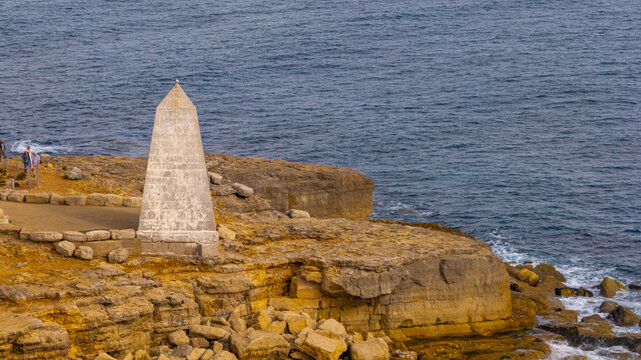 Aerial view of the obelisk monument on the rocky coastline of the Isle of Portland with people walking nearby and the sea in the background, Portland, England, United Kingdom.