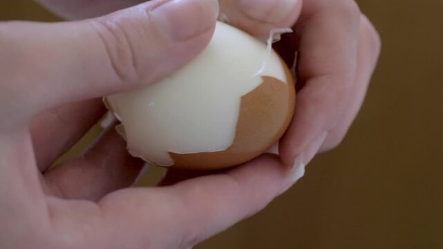 Human hands remove brown shell from hard boiled egg. White surface appears under cracked eggshell. Healthy breakfast recipe, meal prep guide and nutrition blog