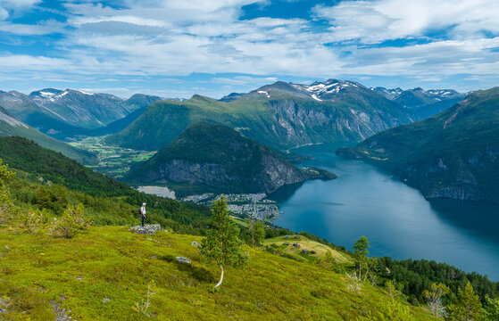 Aerial view of a hiker standing on the grassy slopes of Vardebu overlooking the deep blue waters of Norddalsfjorden, surrounded by mountains and the town of Valldal, More og Romsdal, Norway.