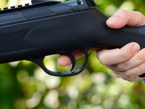 A close-up shot of person's hand holding a black rifle with their index finger resting on the trigger against a green nature background.