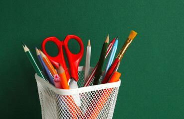 Metal pencil holder filled with colorful school supplies on green surface © Sunlight