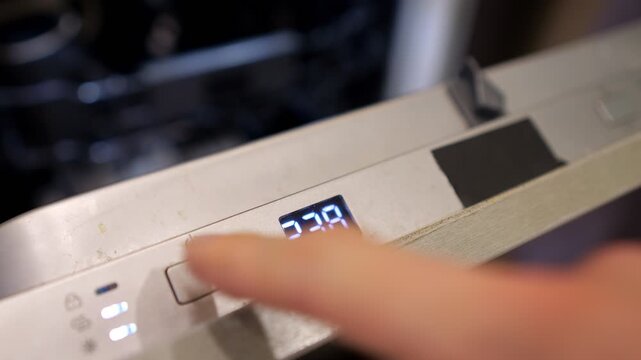 Hand Presses Power Switch On Modern Dishwasher Control Panel Inside Kitchen. Digital Timer Illuminates With Illuminated Numbers, Displaying Cycle Duration During Daily Household Chores.