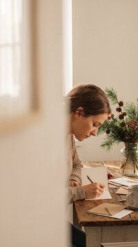 A Woman Writing a Holiday Card: A young woman with her hair tied back sits at a wooden table, diligently writing on a holiday card.