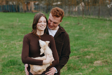 Fototapeta premium A young couple embraces outdoors while the woman holds a small goat in her arms, sharing a tender moment in a casual lifestyle setting.