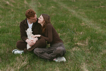 Fototapeta premium Couple sitting on grass in a park, sharing a tender kiss while holding a small white dog, a candid moment of affection and companionship in a natural setting with warm daylight