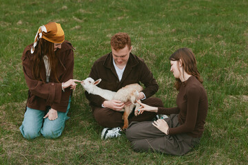 Fototapeta premium Lifestyle scene shows a family in a grassy park with a playful goat kid, three people gently interacting, feeding, and bonding, capturing everyday farm charm and curiosity, a moment of calm, care,