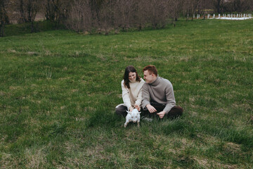 Fototapeta premium Couple with a white goat kid in a grassy field, sharing a gentle moment. Sunlit rural scene conveys lifestyle and affection, natural joy, and relaxed, candid connection between people and animals.