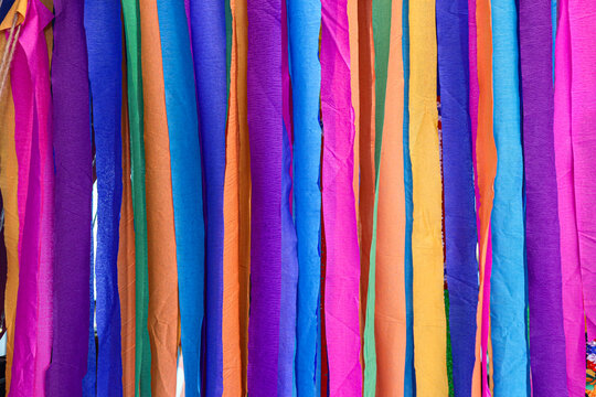 Seven colored offering cloth strips hang at a Thai shrine, showing thin wrinkled textile in vivid vertical bands for spiritual culture articles, travel publishing and seasonal design backgrounds.