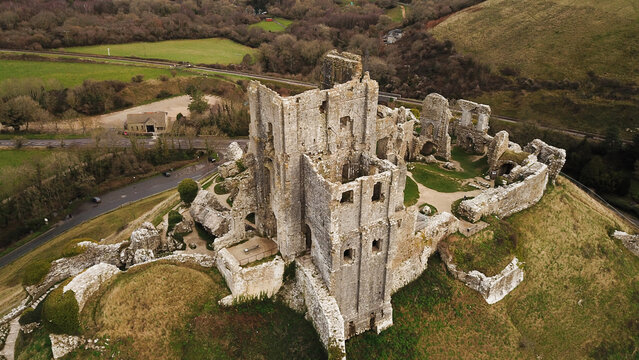 Aerial view of the historic ruins of Corfe Castle set on a grassy hill, surrounded by rolling countryside, roads, and trees in Corfe Castle, England, United Kingdom.