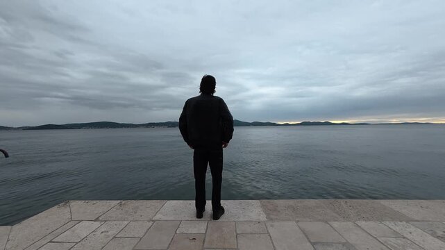 Solitary man reaches stone waterfront terrace to enjoy peaceful horizon gazing. Thoughtful observer admires distant seascape under cloudy twilight, feeling tranquil beside gentle water expanse.