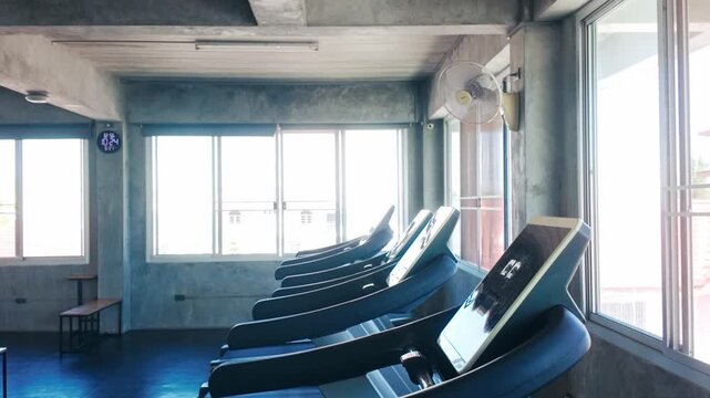 Row of treadmills in a bright, modern gym by large windows, empty and ready for cardio workouts
