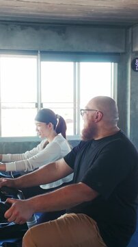 Two people ride stationary bikes in a sunlit gym, smiling and talking during an indoor cycling workout.