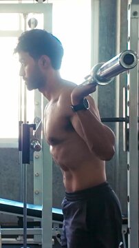 Side-profile of a man performing a barbell back squat in a gym, focused on form and strength training beside a weight rack and window light
